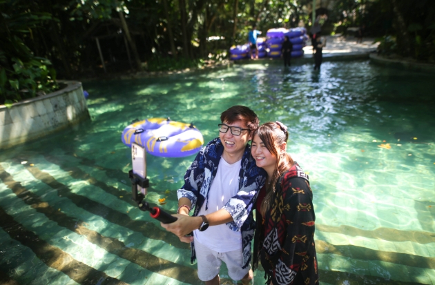 John dan Riana melakukan daily vlog dengan latar belakang Lazy River, salah satu wahana air Bugis Waterpark Adventure.
