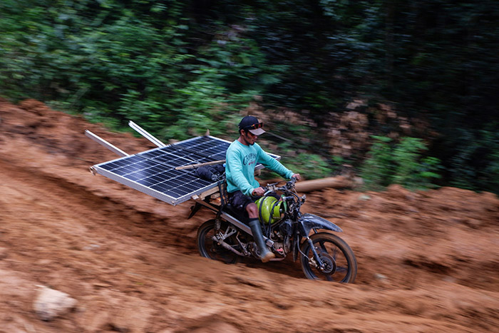 Seorang pengemudi ojek modifikasi mengangkut panel surya SuperSUN melintasi jalur tanah berlumpur.