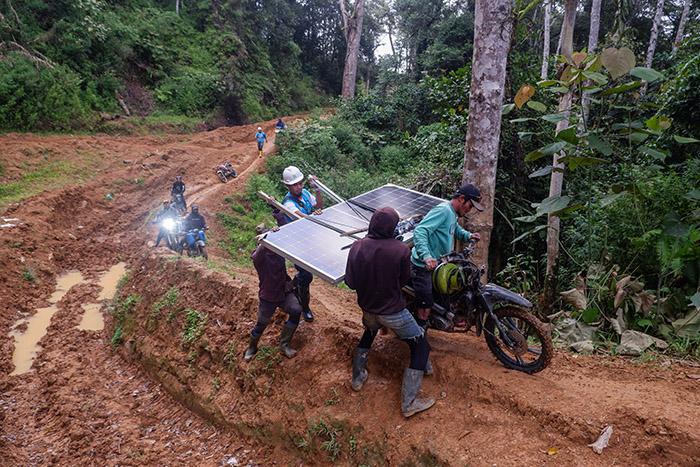 Tim PLN membantu pengemudi ojek modifikasi yang mengangkut panel surya SuperSUN menaklukkan tanjakan berlumpur.