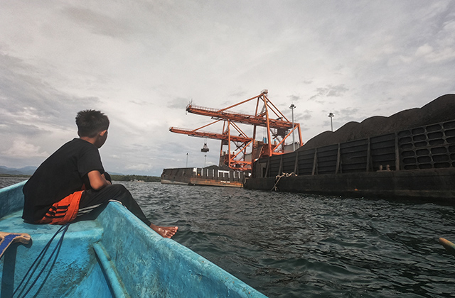 Seorang warga melintas dengan perahu di sekitar aktivitas bongkar muat batu bara di pelabuhan. Batu bara tersebut digunakan untuk memenuhi kebutuhan pembangkit listrik nasional.