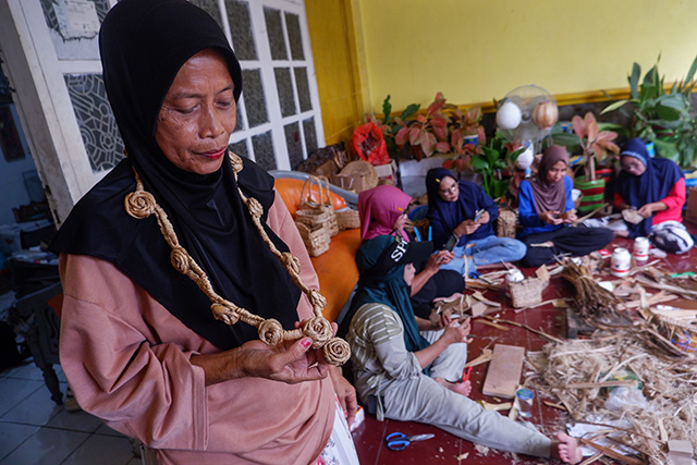 Seorang perajin menunjukkan hasil rangkaian eceng gondok yang diolah menjadi kerajinan, sementara anggota komunitas lainnya tetap sibuk berkarya di ruang produksi sederhana.